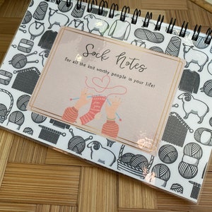 May include: A white notebook with a black spiral binding. The cover features a black and white pattern of yarn balls, knitting needles, and sheep. The front cover has a pink and white sticker with the text "Sock Notes for all the knit worthy people in your life!" and an illustration of two hands knitting.
