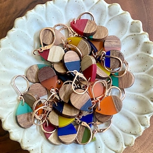 May include: A collection of colorful resin and wood earrings in various shapes and sizes. The earrings are a mix of geometric and organic shapes, with some featuring a single color and others featuring multiple colors. The earrings are all handmade and unique.