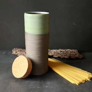 Op de afbeelding: Een keramische pot met een groene en bruine glazuur, een kurken deksel en een bundel spaghetti ervoor.