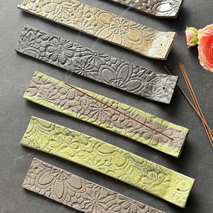 Könnte beinhalten: Sechs Keramik-Räucherstäbchenhalter in verschiedenen Farben, darunter Grau, Beige und Grün, jeweils mit einem geprägten Blumendesign. Räucherstäbchen sind auf den Haltern platziert. Eine rosa Blume und ein dunkler Hintergrund vervollständigen die Szene.