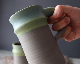 Puede incluir: Una taza de cerámica con un esmalte verde claro y azul en la mitad superior y un esmalte marrón en la mitad inferior. La taza tiene un asa y está siendo sostenida por una mano.