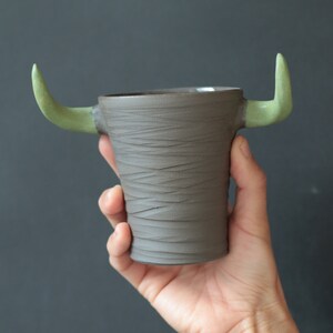 Peut inclure: Une tasse en céramique brune avec des cornes vertes. La tasse a une surface texturée et est tenue dans la main d'une personne.