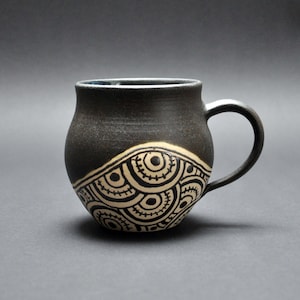 Könnte beinhalten: Eine schwarze Keramiktasse mit einem weißen geometrischen Muster. Die Tasse hat einen abgerundeten Körper und einen Henkel.