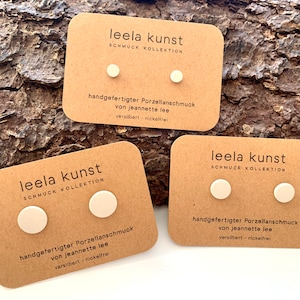 Może przedstawiać: Trzy karty z tekstem "leela kunst" i "SCHMUCK KOLLEKTION". Każda karta ma dwa małe, okrągłe, kremowe kolczyki z porcelany. Tekst na kartach brzmi "handgefertigter Porzellanschmuck von jeannette lee" i "versilbert - nickelfrei".