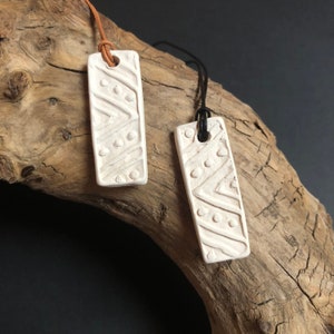 Könnte beinhalten: Zwei cremefarbene rechteckige Anhänger mit geometrischen Mustern. Jeder Anhänger hat oben ein Loch für eine Kordel. Einer hat eine braune Lederschnur, der andere eine schwarze Kordel. Die Anhänger werden auf einem Stück Treibholz präsentiert.