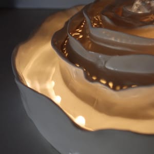 May include: A white ceramic rose-shaped decorative bowl with a warm, glowing interior. The bowl has multiple layers of petals, creating a textured and detailed design. The light source is within the bowl, illuminating the petals.