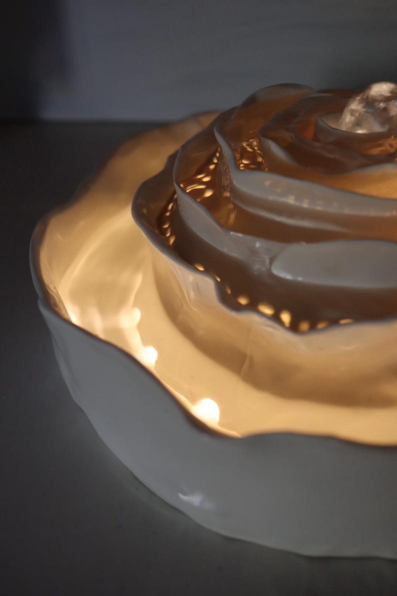 May include: A white ceramic rose-shaped decorative bowl with a warm, glowing interior. The bowl has multiple layers of petals, creating a textured and detailed design. The light source is within the bowl, illuminating the petals.
