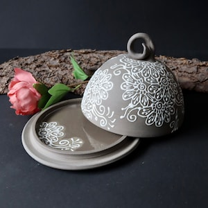 Könnte beinhalten: Eine Butterschale aus Keramik mit Deckel. Die Schale und der Deckel sind hellbraun mit einem weißen Blumenmuster.