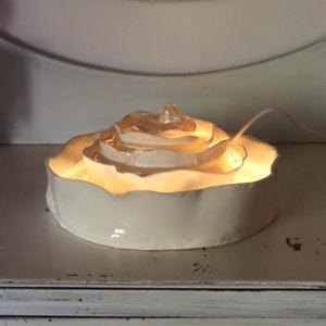 May include: A white ceramic rose-shaped lamp with a warm glow. The lamp has a round base and a detailed, layered design resembling a rose. A clear, decorative element sits atop the rose, and a white cord extends from the back.