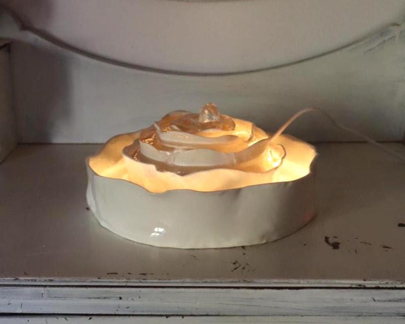 May include: A white ceramic rose-shaped lamp with a warm glow. The lamp has a round base and a detailed, layered design resembling a rose. A clear, decorative element sits atop the rose, and a white cord extends from the back.