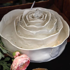 May include: A white ceramic fountain, shaped like a rose, with water cascading from the centre. A pink flower and a brown stone are in the foreground. The design suggests a decorative or garden theme.