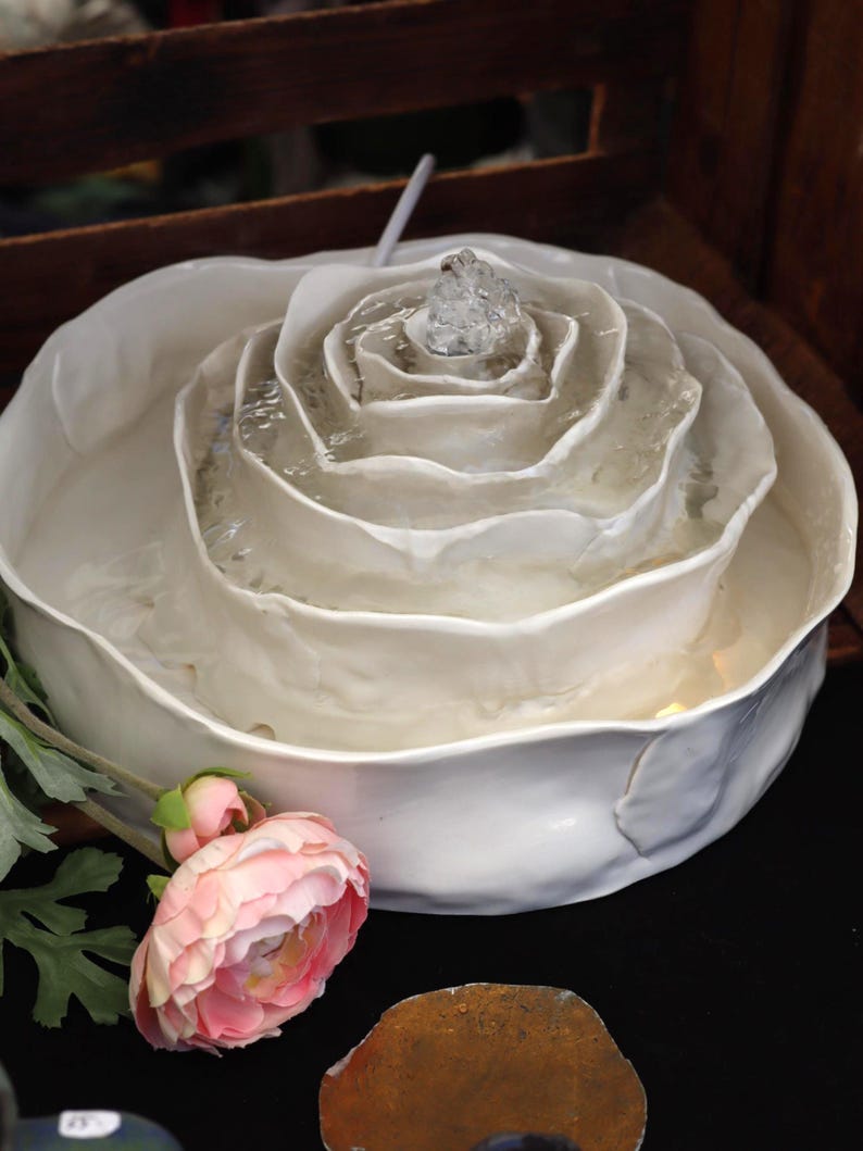 May include: A white ceramic fountain, shaped like a rose, with water cascading from the centre. A pink flower and a brown stone are in the foreground. The design suggests a decorative or garden theme.