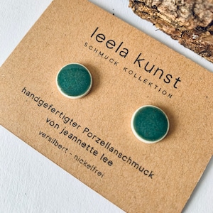Op de afbeelding: Een paar groene keramische oorbel studs op een bruine kaart met de tekst "leela kunst schmuck kollektion handgefertigter porzellan schmuck von jeannette lee versilbert - nickelfrei".