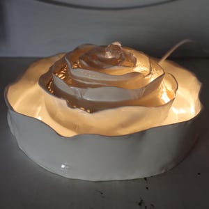 May include: A white ceramic rose-shaped light. The light has a layered petal design with a warm, glowing light. The base is a shallow, scalloped dish. The light is designed to be decorative.