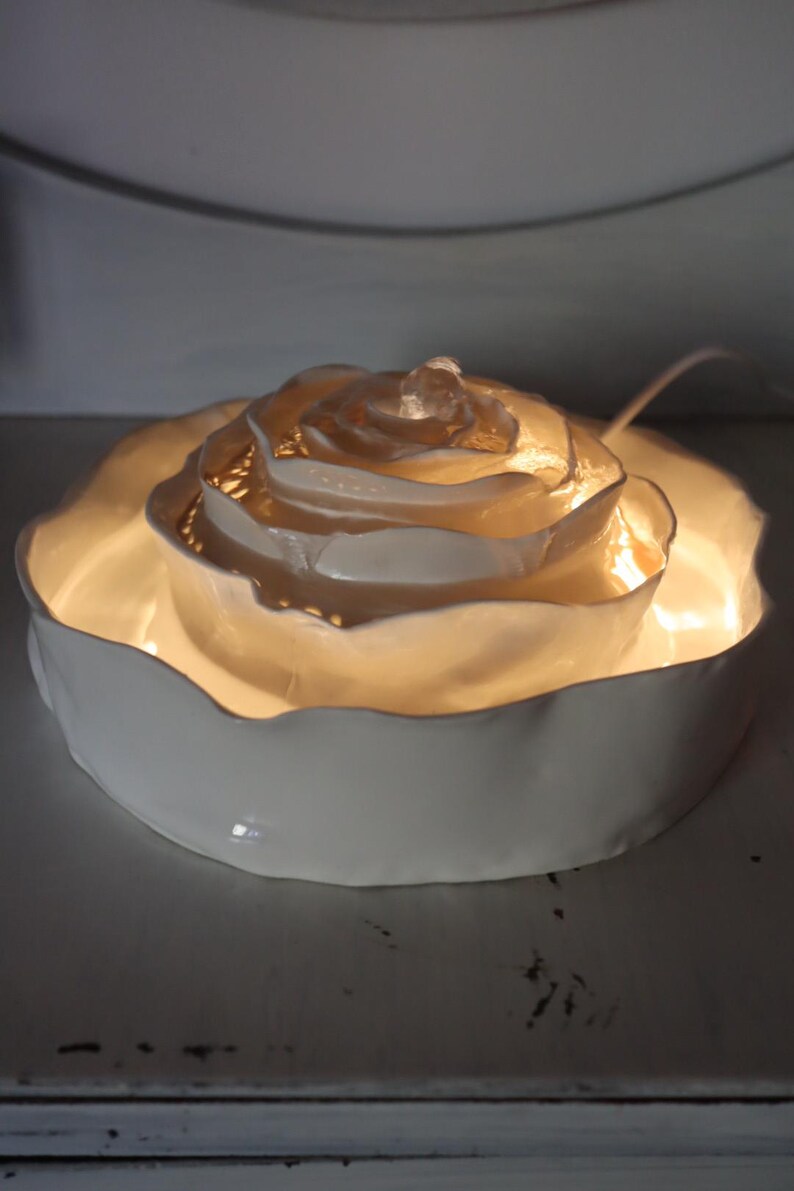 May include: A white ceramic rose-shaped light. The light has a layered petal design with a warm, glowing light. The base is a shallow, scalloped dish. The light is designed to be decorative.