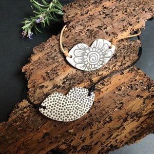 Könnte beinhalten: Zwei herzförmige Keramikanhänger mit Kordelketten. Ein Anhänger hat ein Blumenmuster, der andere ein gepunktetes Muster. Die Anhänger sind weiß mit grauen Designs und schwarzen Kordeln. Der Hintergrund ist ein Stück Holz.