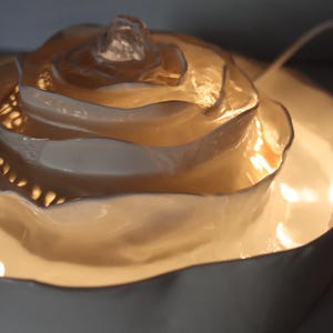 May include: A decorative, illuminated rose-shaped lamp. The lamp has a layered, petal-like design in a creamy white colour, with a central, crystal-like element. The lamp sits on a matching, circular base, and emits a warm, inviting glow.