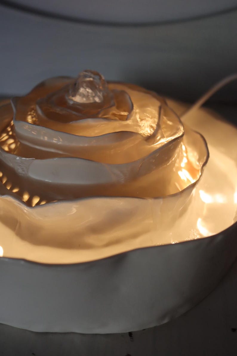 May include: A decorative, illuminated rose-shaped lamp. The lamp has a layered, petal-like design in a creamy white colour, with a central, crystal-like element. The lamp sits on a matching, circular base, and emits a warm, inviting glow.