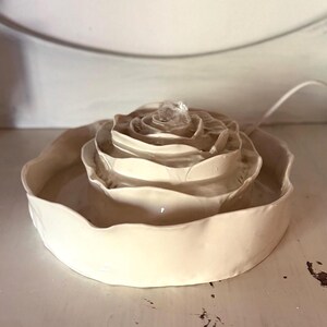 May include: A cream ceramic rose-shaped fountain. The fountain features a layered design, with water gently flowing from the centre. The base is a shallow dish, and a power cord is visible.