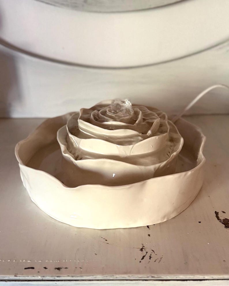 May include: A cream ceramic rose-shaped fountain. The fountain features a layered design, with water gently flowing from the centre. The base is a shallow dish, and a power cord is visible.