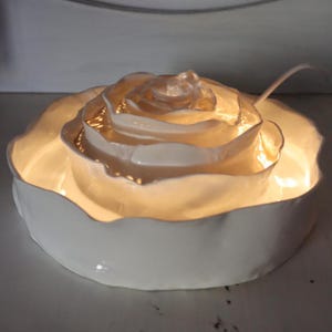 May include: A white ceramic rose-shaped fountain with a soft, glowing light emanating from within. The fountain has multiple layers of petals, creating a delicate and elegant design. The cord is visible.