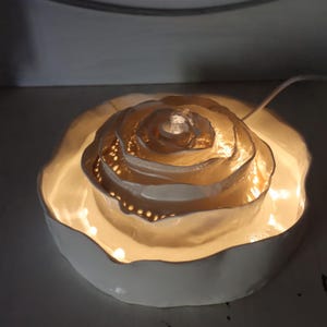 May include: A white ceramic rose-shaped lamp with a warm glow. The lamp has multiple layers, with a central light source and small perforations around the edges. The lamp sits in a shallow, scalloped dish.