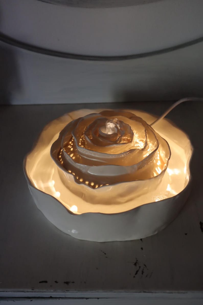 May include: A white ceramic rose-shaped lamp with a warm glow. The lamp has multiple layers, with a central light source and small perforations around the edges. The lamp sits in a shallow, scalloped dish.