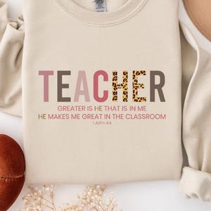 May include: A beige sweatshirt with the words "TEACHER" in pink, brown, and leopard print. The text "GREATER IS HE THAT IS IN ME HE MAKES ME GREAT IN THE CLASSROOM 1 John 4:4" is printed below.