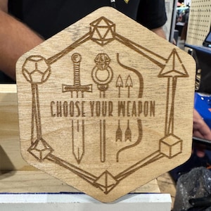 May include: A wooden, hexagonal plaque with laser-etched designs. The plaque features a sword, ring, arrows, and the text "CHOOSE YOUR WEAPON". Geometric shapes frame the design, creating a unique, rustic aesthetic.