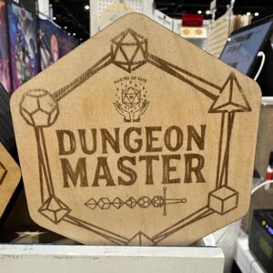 May include: A light brown wooden sign featuring the words "DUNGEON MASTER" in a bold font. The sign has a geometric design with a dice and sword. The text "MASTER OF FATE" is above the main text. The sign is hexagonal.