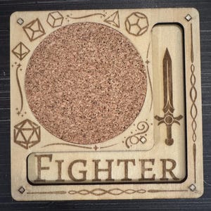 May include: A wooden coaster with a cork center, featuring engraved dice, a sword, and the word "FIGHTER." The coaster has a square shape with rounded corners and decorative etched borders. The cork center is circular.