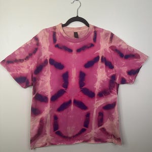 May include: A pink and purple tie-dye t-shirt hanging on a black hanger. The short-sleeved shirt has a symmetrical pattern. The background is a plain white wall. The tie-dye design is in shades of pink and purple.