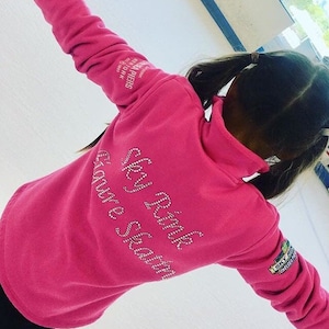 以下が含まれることがあります： 背面にラインストーンで「Sky Rink Figure Skating」と書かれたピンクのトレーナー。左袖には「Sky Rink Chelsea Piers」のロゴがあります。