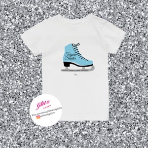 May include: White t-shirt with a blue figure skate graphic and the text "Figure Skater". The text "Graceful, Powerful, Strong, Determined" is printed below the skate. The text "GlitzyUpGifts" is printed on a white circle with a pink outline.