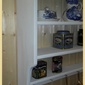 May include: White wooden wall shelf with three shelves and hooks. The shelves are filled with various items, including a blue and white ceramic pitcher, a blue and white ceramic butter dish, two blue and white tea tins, and two colorful tins. Two copper pots hang from the hooks.