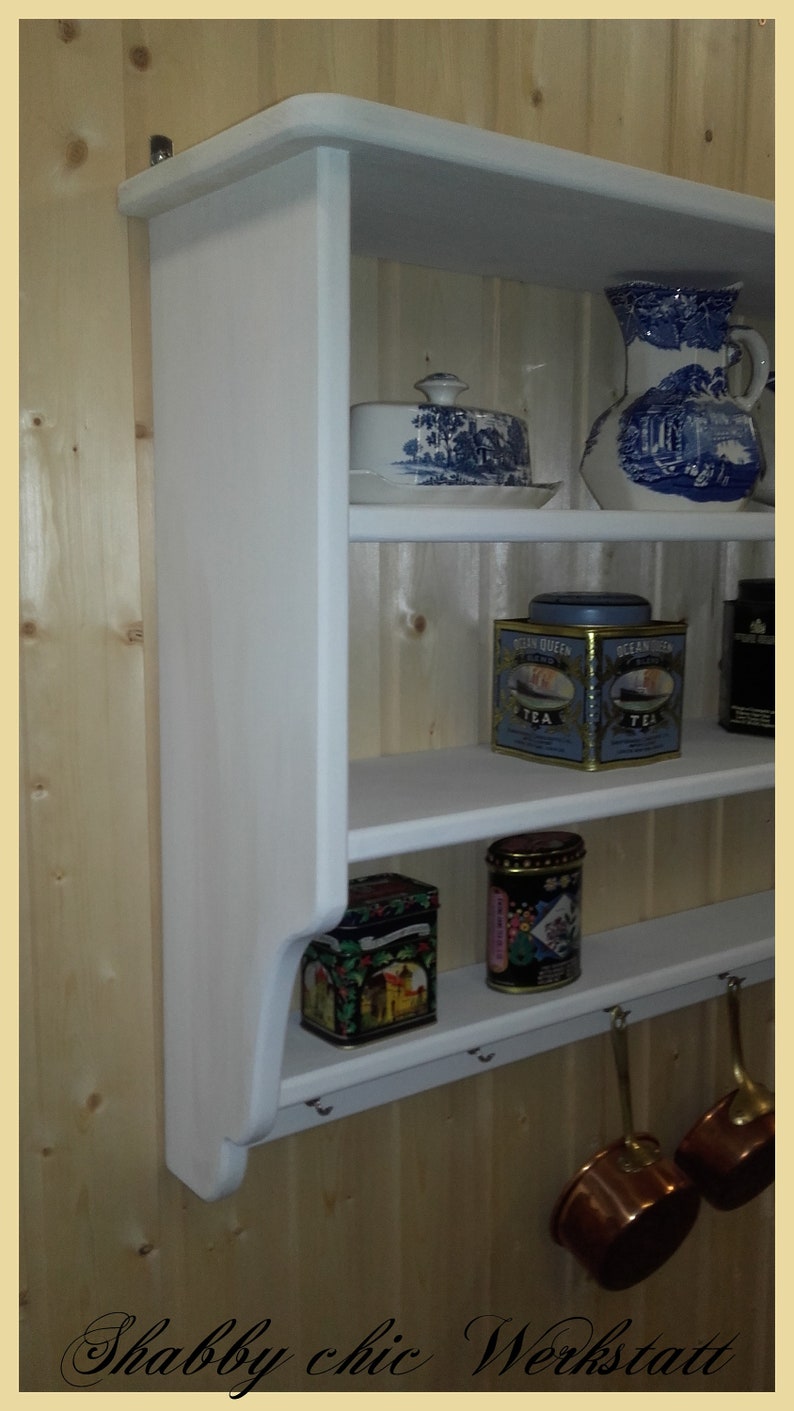 May include: White wooden wall shelf with three shelves and hooks. The shelves are filled with various items, including a blue and white ceramic pitcher, a blue and white ceramic butter dish, two blue and white tea tins, and two colorful tins. Two copper pots hang from the hooks.
