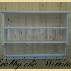 May include: White wooden wall shelf with three shelves and a row of hooks on the bottom. The shelf is mounted on a wood paneled wall. The text "Shabby chic Werkstatt" is written below the shelf.