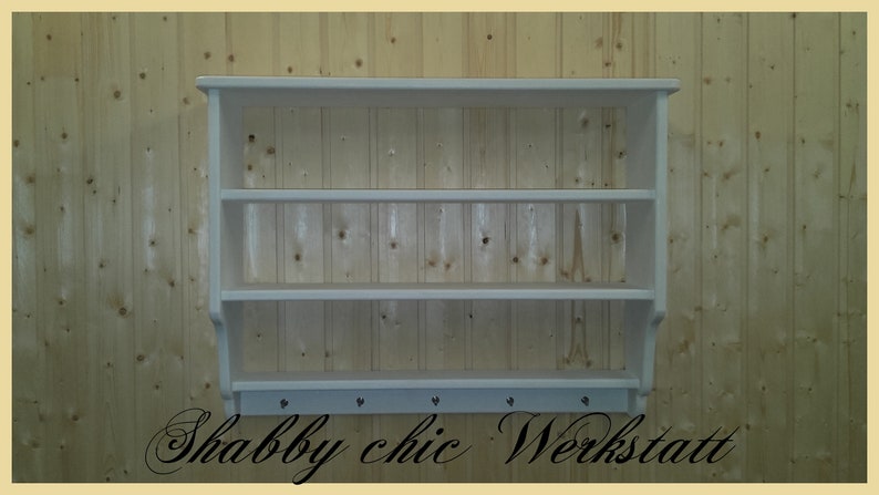 May include: White wooden wall shelf with three shelves and a row of hooks on the bottom. The shelf is mounted on a wood paneled wall. The text "Shabby chic Werkstatt" is written below the shelf.