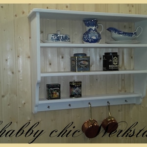 May include: White wooden wall shelf with three shelves and hooks underneath. The shelf is decorated with vintage-style dishes, tea tins, and other items. The text "Shabby chic Werkstatt" is written below the shelf.