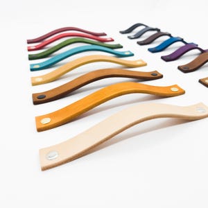 May include: A collection of leather drawer pulls in various colors, including red, green, blue, and brown. Each pull has a curved design and is attached with metal rivets. The pulls are arranged on a white background.