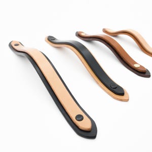 May include: Four leather cabinet pulls in varying colors: natural, black, and two shades of brown. Each pull is curved and features a contrasting edge and a small, dark rivet at each end. The pulls are designed for furniture.