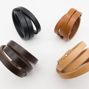 Könnte beinhalten: Vier Lederarmbänder in Schwarz, Braun, Beige und Hellbraun. Jedes Armband besteht aus drei Lederriemen, die sich um das Handgelenk wickeln. Die Armbänder werden auf weißem Hintergrund präsentiert.