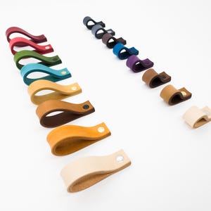 May include: A collection of leather cable organizers in various colors, including red, green, blue, and brown. Each organizer is a curved loop with a metal snap closure. The organizers are arranged on a white surface.