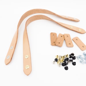 May include: A set of light brown leather bag handles with metal rivets, wooden reinforcement pieces, and various hardware including silver, gold, and black rivets and screws, all on a white background.