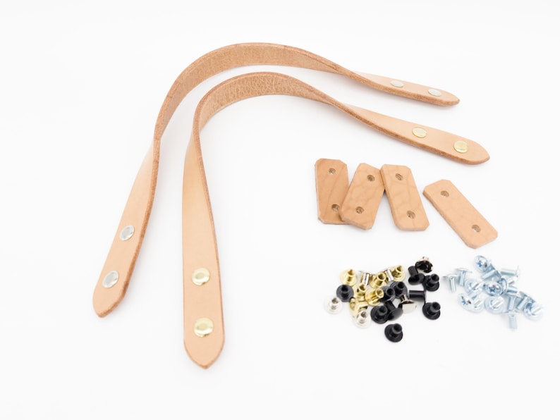 May include: A set of light brown leather bag handles with metal rivets, wooden reinforcement pieces, and various hardware including silver, gold, and black rivets and screws, all on a white background.