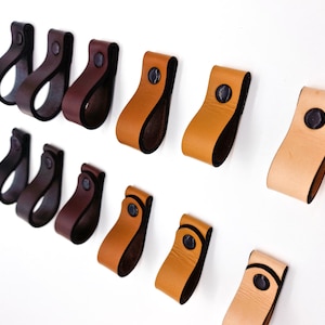 May include: A selection of leather cabinet pulls in shades of black, dark brown, tan, and light brown. Each pull has a loop design and a dark rivet. The pulls are displayed against a white background.