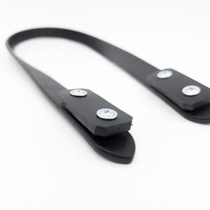 May include: A black leather strap with two rectangular end pieces, each secured with two silver screws. The strap curves gently against a white background, suggesting a handle or attachment for a bag or similar item.