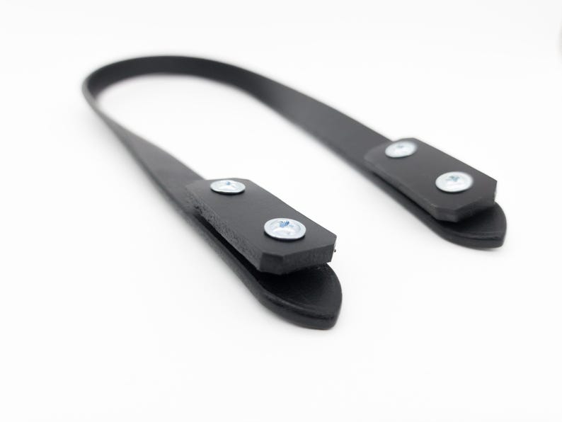 May include: A black leather strap with two rectangular end pieces, each secured with two silver screws. The strap curves gently against a white background, suggesting a handle or attachment for a bag or similar item.