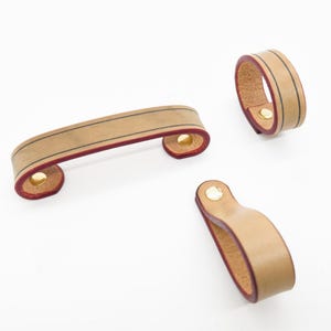 Könnte beinhalten: Eine Sammlung von Leder-Schrankgriffen und -zügen in hellbrauner Farbe. Die Griffe haben eine dunkelrote Kante und einen dünnen schwarzen Streifen. Die Hardware ist mit goldfarbenen Nieten befestigt. Das Set enthält einen langen Griff, einen ringförmigen Griff und einen kleinen Schlaufengriff.