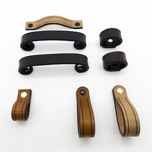 Può includere: Un assortimento di ferramenta per mobili in pelle in vari stili e colori. Include maniglie curve in nero, marrone e beige, insieme a tiranti e pomelli circolari abbinati. Ferramenta per mobili e armadi.